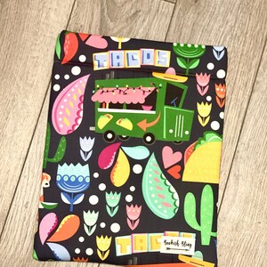 May include: A black e-reader cover with a vibrant taco truck pattern. The design includes the word "TACOS", a green truck, tacos, flowers, and cacti. The words "Bookish Bling" are also visible on the cover.