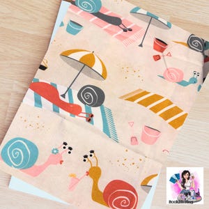 May include: A light beige fabric with a beach-themed pattern. The design features cartoon snails relaxing on striped beach towels under umbrellas. Buckets, shovels, and drinks are also included in the illustration.