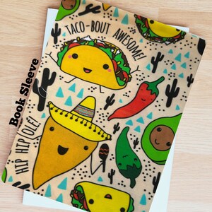 May include: A tan book sleeve with a repeating pattern of cartoon tacos, avocados, chili peppers, and cacti. The text "TACO-BOUT AWESOME!" and "HIP HIP OLE!" are included. The words "Book Sleeve" are printed vertically on the left side.