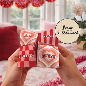 Puede incluir: Una caja de regalo de múltiples lados con las palabras "LOVE", "XOXO", "FOREVER" y "FRIEND" impresas en los lados. La caja tiene un diseño en forma de corazón en tonos de rosa, naranja y blanco. La caja se sostiene frente a un fondo borroso.