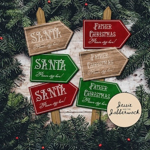 May include: Six wooden arrow signs with white text on a whitewashed wood background. The signs say "Santa" and "Father Christmas" with the text "Please stop here!" on each sign. The signs are surrounded by green pine branches with red berries.