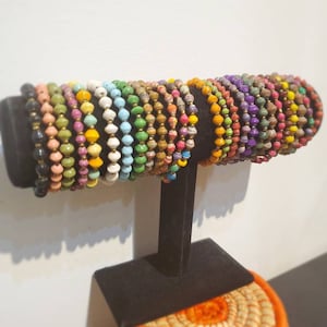 May include: A display of colorful beaded bracelets made from recycled paper. The bracelets are stacked on a black stand and are arranged in a rainbow pattern.