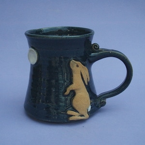 May include: A dark teal ceramic mug with a tan rabbit applique and a white moon. The mug has a large handle and a flared rim. The rabbit is standing on its hind legs, looking up. The mug is approximately 10 cm tall.