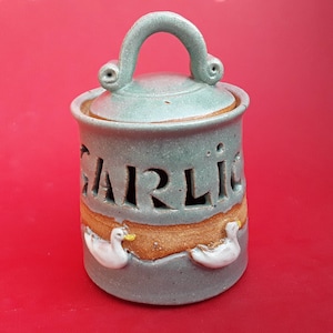 May include: A ceramic garlic keeper with a lid and handle. The word "GARLIC" is cut out of the grey-blue container. Two white duck figures are on the lower part. The container sits on a red surface.