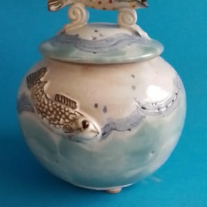 May include: A ceramic jar with a lid, featuring a fish design. The jar is primarily off-white with blue wave and fish accents. A fish figurine adorns the lid. The jar is round and has a small, round base. The background is a solid blue.