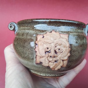 May include: A small brown ceramic bowl with two handles and a green face sculpted into the side.