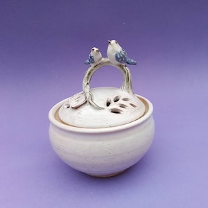 May include: White ceramic jar with a lid featuring a twig handle with two small blue birds perched on top. The lid has a decorative design of leaves and holes.