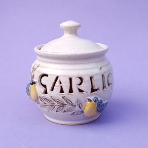 May include: Cream ceramic garlic pot with a lid. The word "GARLIC" is cut out of the jar. Two small, colourful birds are perched on the jar, with olive branch details. The jar is on a light purple background.