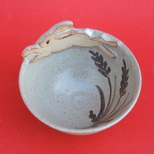 May include: A small, speckled ceramic bowl with a leaping rabbit figurine on the rim. The bowl features brown wheat stalk designs and is set against a red background. A charming, handcrafted piece.