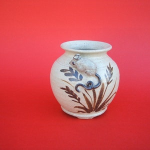 Könnte beinhalten: Eine weiße Keramikvase mit einem braunen Maus-Design und braunen, bemalten Weizenähren. Die Vase hat einen runden Körper und eine leicht ausgestellte Öffnung.