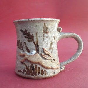 May include: A ceramic mug with a brown and white glaze features a brown rabbit jumping through tall grass. The mug has a brown handle.
