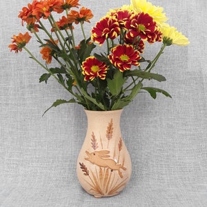 May include: A ceramic vase featuring a brown rabbit design and wheat stalks. The vase is filled with a bouquet of orange, red and yellow chrysanthemums.