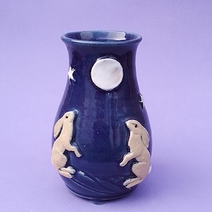 Könnte beinhalten: Eine dunkelblaue Keramikvase mit einem Nachthimmel-Thema. Die Vase zeigt zwei hellbraune Hasen, einen weißen Mond und weiße Sterne. Die Vase hat eine glänzende Oberfläche und eine abgerundete Form, geeignet für Blumen.