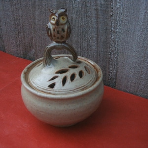 May include: A ceramic jar with a lid featuring a brown and white owl figurine. The lid has a leaf design with cut-out holes.