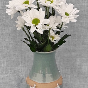 May include: A bouquet of white daisies in a light blue ceramic vase with a brown band and white duck figurines around the rim.