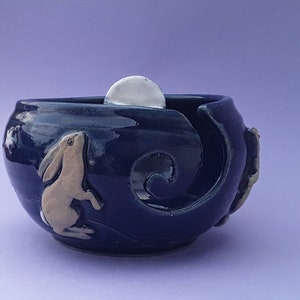 May include: A dark blue ceramic yarn bowl with a glossy finish. The bowl features a raised design of a rabbit looking up at a moon. The moon is a white sphere. The bowl has a swirl design on the side.
