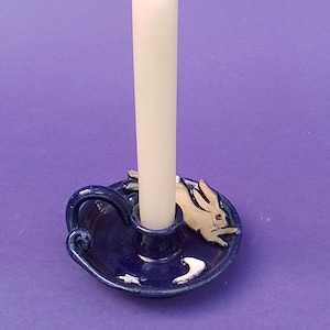 May include: A blue ceramic candle holder with a white candle. The holder has a rabbit design and a handle. The candle is white and stands upright. The holder is on a purple background.