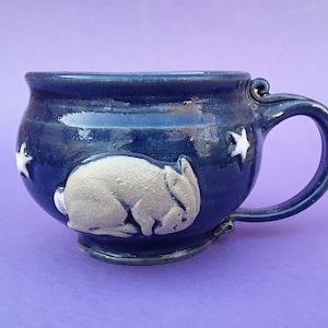 May include: A blue ceramic mug with a white rabbit design and white stars. The mug has a rounded body and a handle.