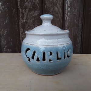 May include: A ceramic garlic keeper with a lid. The jar is light blue and white with the word "GARLIC" cut out of the side. The keeper is round with a small handle on the lid.