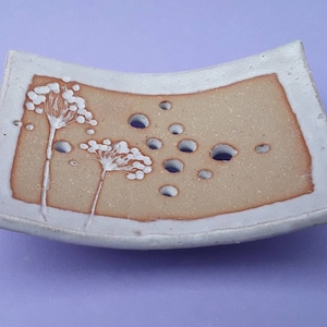 May include: A handmade ceramic soap dish with a light brown centre and white edges. The dish features a floral design and several drainage holes. The shape is rectangular with slightly curved sides.