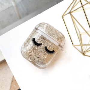 May include: A clear plastic case for AirPods with gold glitter and black eyelashes printed on the front.