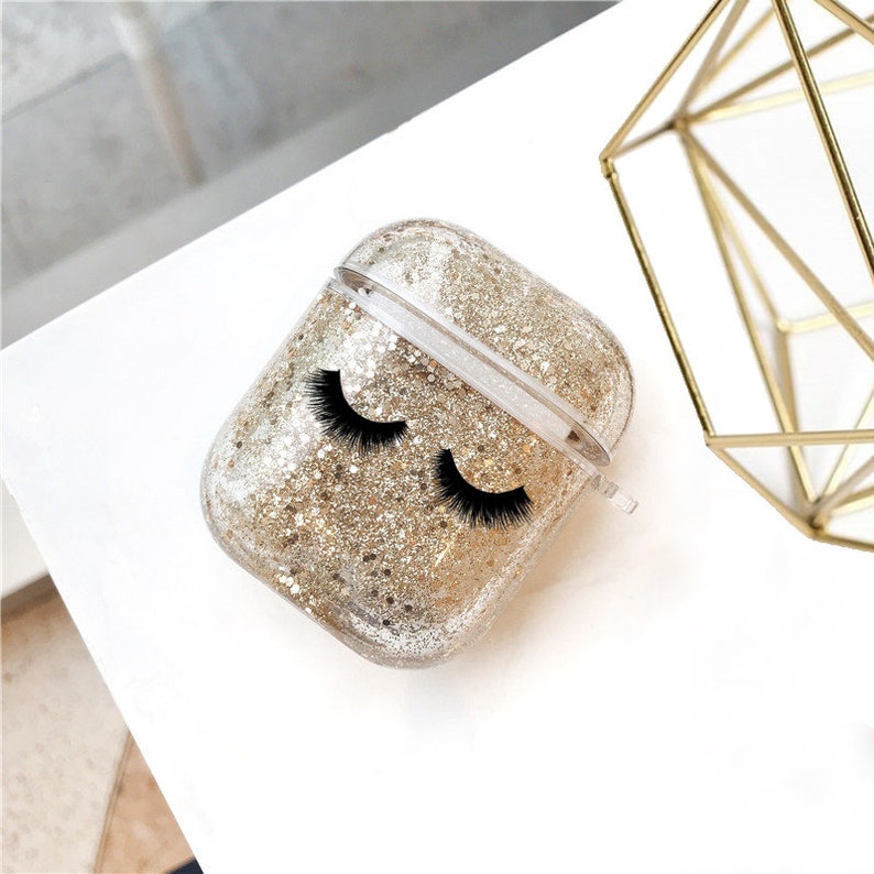 May include: A clear plastic case for AirPods with gold glitter and black eyelashes printed on the front.