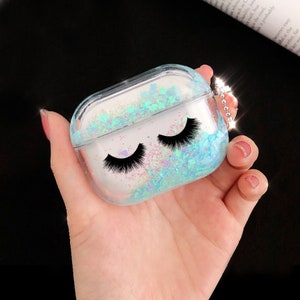 May include: Clear plastic case for AirPods with a sparkly blue and white glitter design and black eyelashes. The case has a small silver chain attached.