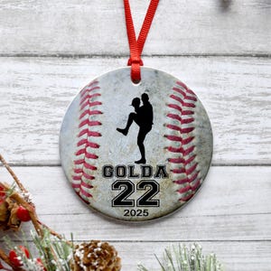 May include: A white and red baseball ornament with a black silhouette of a baseball player pitching. The ornament has the name "GOLDA" and the number "22" printed on it, along with the year "2025".