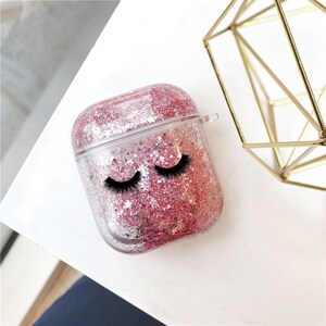 May include: A clear plastic case for AirPods with a pink and silver glitter design. The case has a pair of black eyelashes printed on the front.