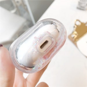 May include: Clear plastic case for wireless earbuds with a glittery pink and white design. The case has a charging port on the bottom.