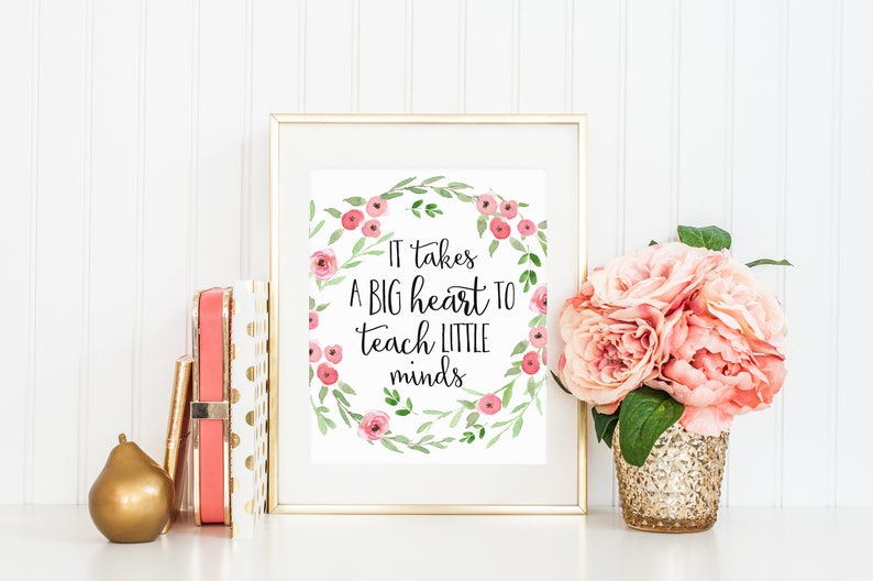May include: A framed print with a floral wreath and the text "It takes a big heart to teach little minds". The print is on a white background and is framed in a gold frame. The print is sitting on a white surface with a gold pear and a stack of books.