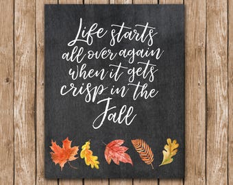 May include: Black chalkboard sign with white text that reads "Life starts all over again when it gets crisp in the fall". The sign is decorated with five watercolor autumn leaves in shades of red, orange, and yellow.