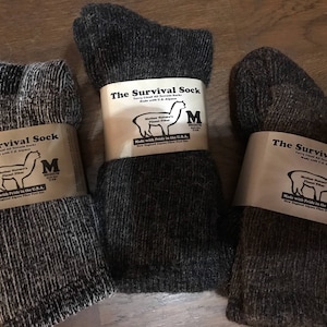Può includere: Tre paia di calze in lana di alpaca grigio scuro con etichette bianche che recitano "The Survival Sock" e presentano un'illustrazione di alpaca. Le etichette includono anche il testo "Mother Nature's Finest Fiber" e "Made with Pride in the U.S.A."