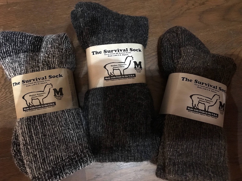 May include: Three pairs of dark gray alpaca wool socks with white labels that read "The Survival Sock" and feature an alpaca illustration. The labels also include the text "Mother Nature's Finest Fiber" and "Made with Pride in the U.S.A."