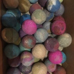 May include: A box filled with colorful felt balls. The balls are various shades of blue, green, purple, pink, and brown. They are all about the size of a golf ball.