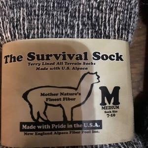 May include: A pair of black and white alpaca socks with a brown label that reads "The Survival Sock" and "Terry Lined All Terrain Socks Made with U.S. Alpaca". The label also features an alpaca illustration and the text "Mother Nature's Finest Fiber" and "Made with Pride in the U.S.A. New England Alpaca Fiber Pool Inc.". The label also includes a size tag that reads "M MEDIUM Sock Size 7-10".