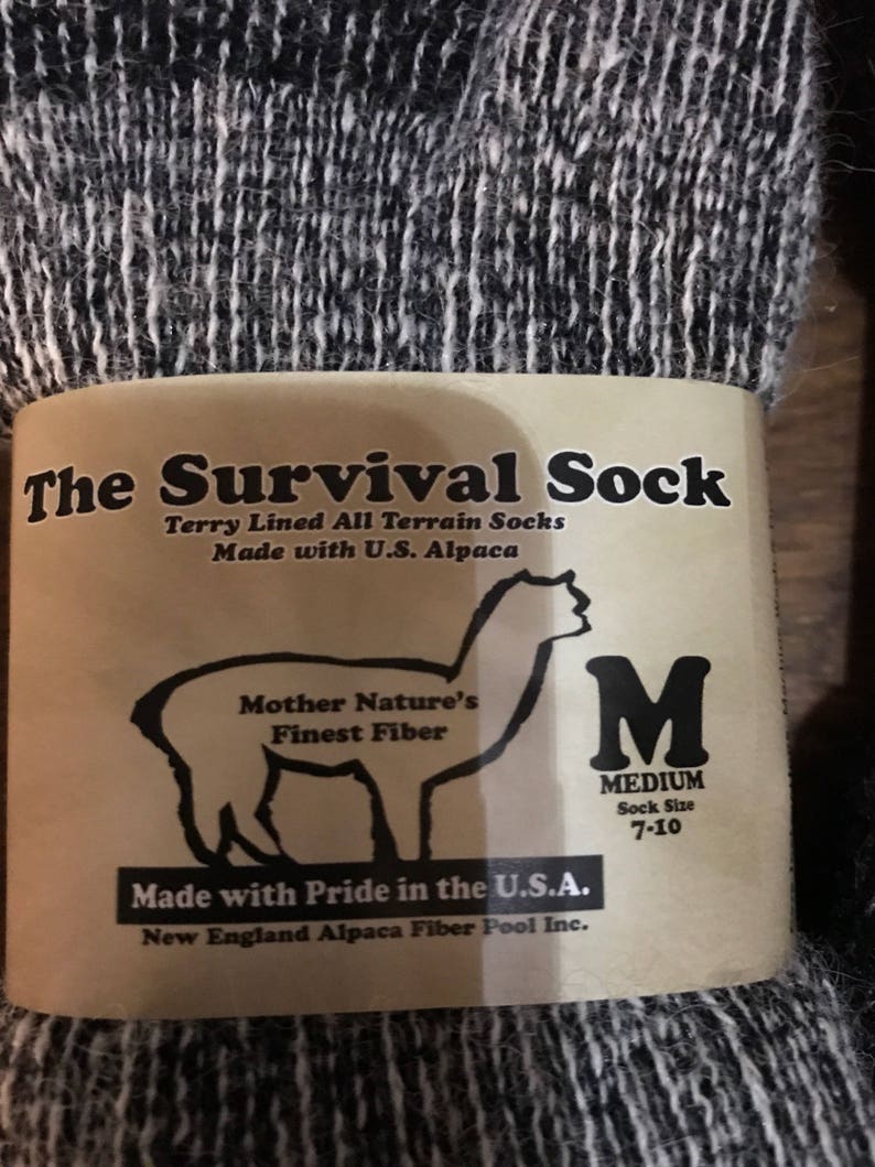 May include: A pair of black and white alpaca socks with a brown label that reads "The Survival Sock" and "Terry Lined All Terrain Socks Made with U.S. Alpaca". The label also features an alpaca illustration and the text "Mother Nature's Finest Fiber" and "Made with Pride in the U.S.A. New England Alpaca Fiber Pool Inc.". The label also includes a size tag that reads "M MEDIUM Sock Size 7-10".