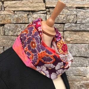 Peut inclure: Une écharpe infinie à motifs floraux colorés avec un schéma de couleurs rose, orange et violet. L'écharpe est en tissu et est exposée sur un mannequin en bois.