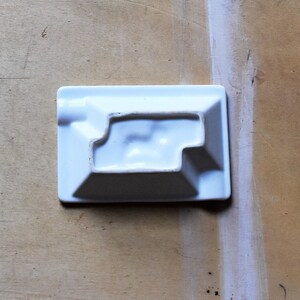 May include: A rectangular, white ceramic ashtray with a stepped interior design. The ashtray has a small, rounded notch on one side for resting a cigarette. The object is set against a light brown wooden surface.