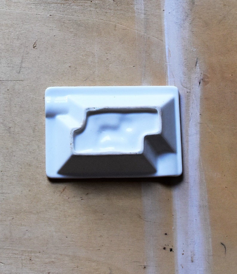 May include: A rectangular, white ceramic ashtray with a stepped interior design. The ashtray has a small, rounded notch on one side for resting a cigarette. The object is set against a light brown wooden surface.
