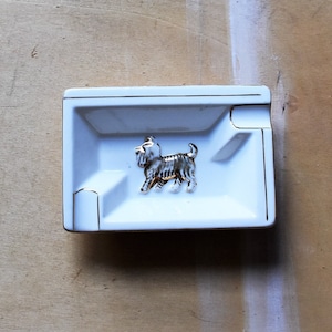 May include: A white rectangular ashtray with gold trim and a decorative terrier figurine in the center. The ashtray has a small indentation for resting cigarettes. The dog figurine is silver and gold.