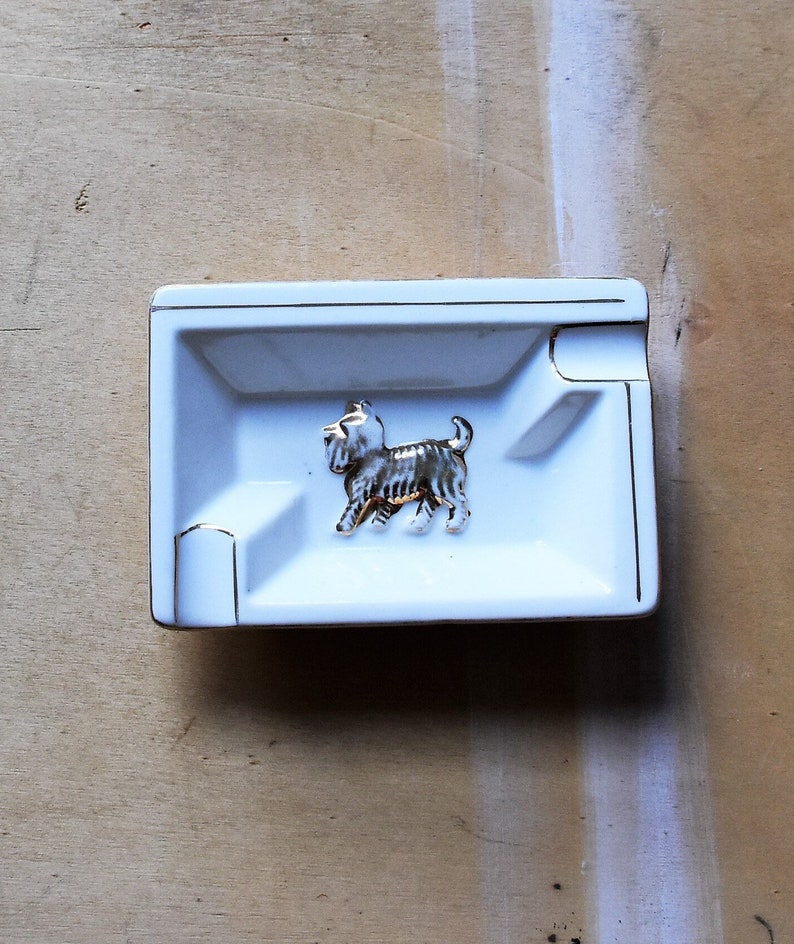May include: A white rectangular ashtray with gold trim and a decorative terrier figurine in the center. The ashtray has a small indentation for resting cigarettes. The dog figurine is silver and gold.