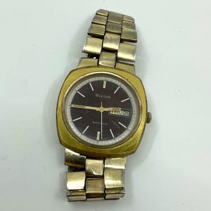 May include: A gold-toned Bulova automatic wristwatch with a brown face and a gold-toned metal strap. The watch face has a date window that displays "THU 26".