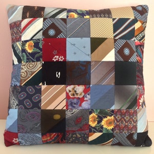 May include: Square decorative pillow with a patchwork design made from various patterned neckties. The pillow features a mix of colors, including blue, red, brown, and floral patterns. The pillow is approximately 16 inches square.