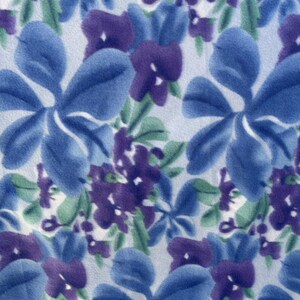 May include: A repeating floral pattern with large blue flowers, smaller purple flowers, and green leaves on a white background. The design has a soft, textured appearance, creating a delicate and natural aesthetic.