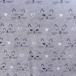 May include: A gray fabric with a repeating pattern of cat faces and white hearts. The cat faces are drawn with black lines, featuring closed eyes, whiskers, and small pink noses. The hearts are scattered throughout the design.