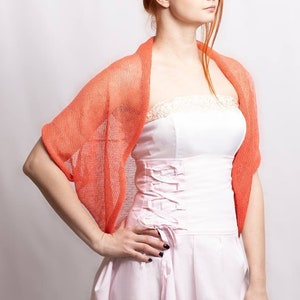 May include: A coral-colored, open-knit shrug with a lightweight, semi-sheer texture worn over a white strapless dress featuring a lace-trimmed neckline and a corset-style bodice with side lacing details and a pleated skirt.