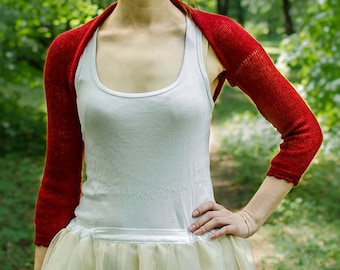 May include: A bright red knitted shrug with three-quarter length sleeves and a scalloped edge, worn over a plain white tank top and a light-colored skirt. The garment features a fine, lightweight knit texture and a simple open-front design.