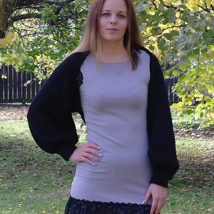 May include: A woman wearing a gray dress with a black lace trim and a black knitted shrug with long sleeves.