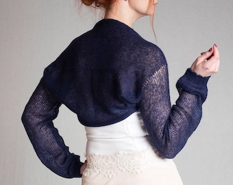May include: A dark navy blue knit bolero shrug featuring long sleeves with ribbed cuffs and a lightweight, semi-sheer open weave texture. The garment is cropped, ending just above the waist, and is worn over a white bodice with lace trim.